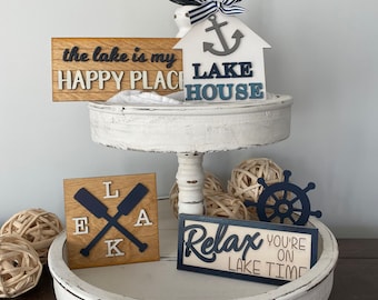 May include: A white tiered tray with a variety of wooden signs with lake-themed sayings. The signs are decorated with blue and white nautical accents, including an anchor, a steering wheel, and oars. The top sign reads "the lake is my happy place LAKE HOUSE". The bottom sign reads "LAKE LIFE 'cause beaches be salty".
