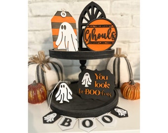 May include: A tiered tray decorated with Halloween-themed decorations. The top tier features a black and orange sign that reads "All the Scary Ghouls are Sweeping Thru". The middle tier features a white ghost cutout on a black tag and a black and orange sign that reads "You look la BOO-lous". The bottom tier features a black and white banner that reads "BOO" and a white ghost cutout.