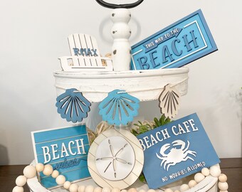 May include: A white tiered tray decorated with a beach theme. The top tier features a sign that says "This Way to the Beach" and a miniature beach chair with the word "Relax" on it. The bottom tier has a sign that says "Beach Vibes", a sign that says "Beach Cafe No Worries Allowed", a sand dollar, and a string of wooden beads.