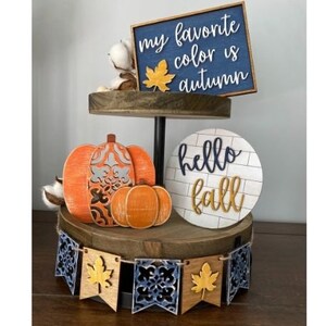 May include: A tiered wooden tray decorated with fall-themed decorations. The top tier features a blue and white sign that reads "my favorite color is autumn". The middle tier features a white sign with black lettering that reads "hello fall". The bottom tier features a pumpkin and a smaller pumpkin with a banner that reads "hello fall".