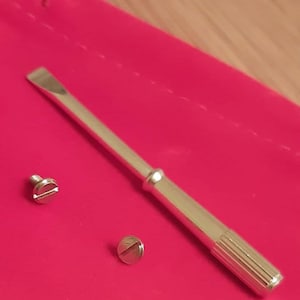 May include: A silver metal tool with a flat head and a ridged handle, along with two small screws, rests on a bright pink surface. The tool appears to be a specialized screwdriver or prying instrument, likely for precision work.