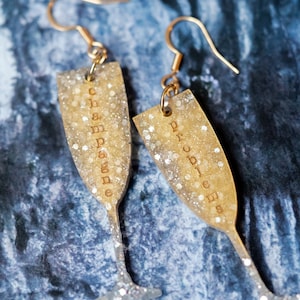 Peut inclure: Une paire de boucles d'oreilles en forme de flûte à champagne en paillettes dorées. La boucle d'oreille gauche dit "Champagne" et la boucle d'oreille droite dit "Cheers".