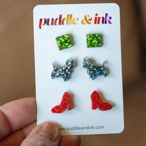 May include: A card with the text "puddle & ink" and the website address "www.puddleandink.com". The card features three pairs of earrings: green glitter squares, black glitter Scottie dogs, and red glitter high heels.