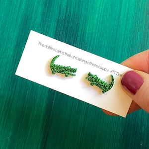 May include: A pair of green glitter alligator earrings. The earrings are on a white card with the text "The noblest art is that of making others happy. - PT Bar"