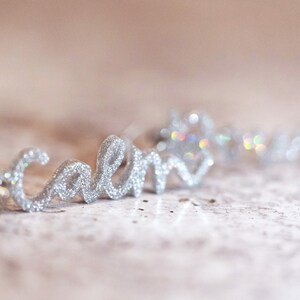 May include: Silver glitter word "calm" in cursive script, with a wire loop at the end. The word is on a light beige surface.