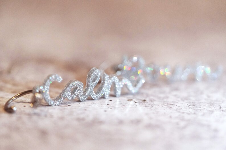 May include: Silver glitter word "calm" in cursive script, with a wire loop at the end. The word is on a light beige surface.