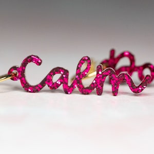 May include: A pair of pink glitter earrings with the word "calm" on one earring and "down" on the other.