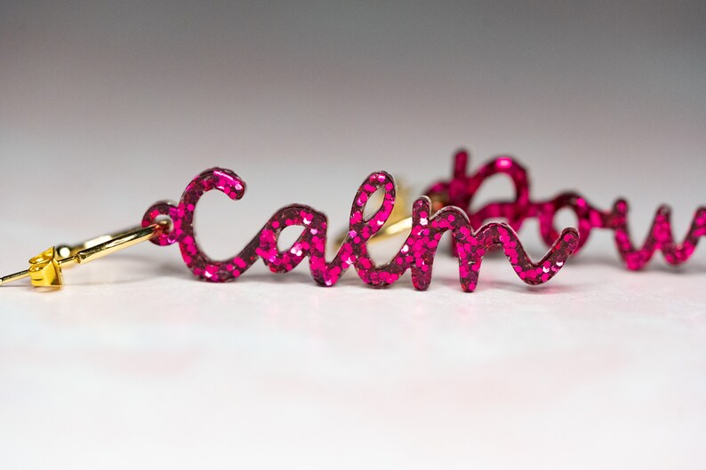May include: A pair of pink glitter earrings with the word "calm" on one earring and "down" on the other.