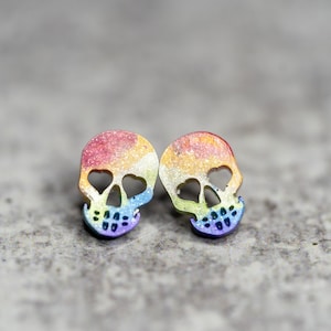 May include: A pair of rainbow glitter skull earrings. The skulls are outlined in black and have heart-shaped eye sockets.