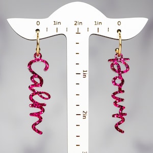 May include: A pair of pink glitter earrings with the words "Calm" and "Down" written in cursive. The earrings are hanging from gold hoops.