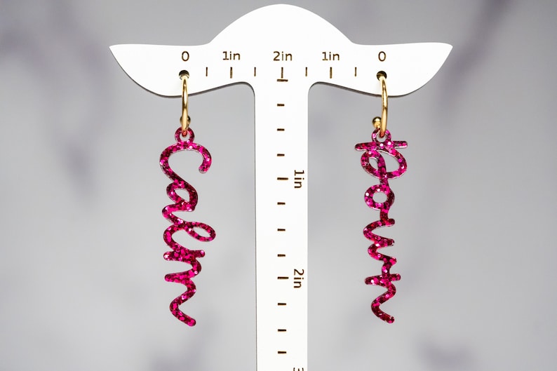 May include: A pair of pink glitter earrings with the words "Calm" and "Down" written in cursive. The earrings are hanging from gold hoops.