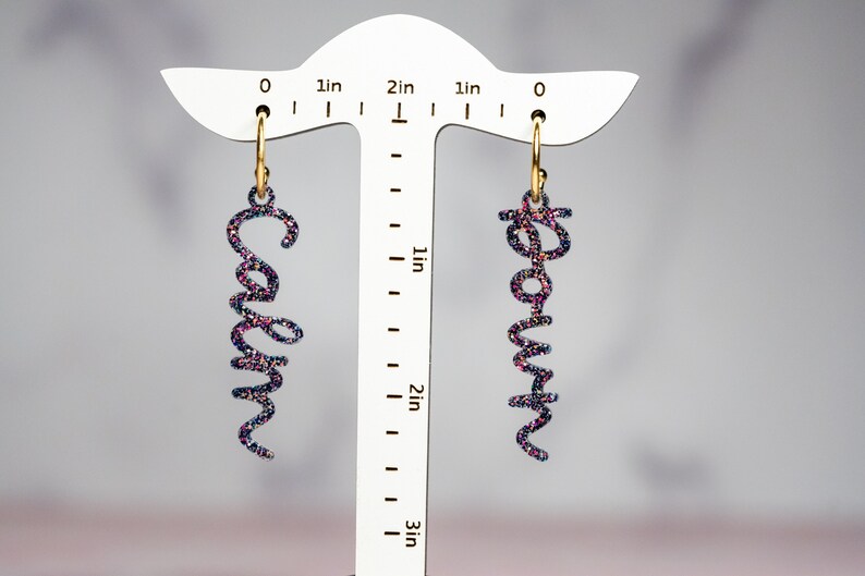 May include: A pair of gold hoop earrings with a glittery black and pink name charm hanging from each hoop. The charms spell out the names "Carolyn" and "Rowan".