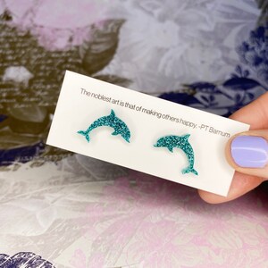 May include: Teal glitter dolphin earrings displayed on a white card. The card features the quote "The noblest art is that of making others happy." -PT Barnum. The background has a floral design.