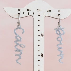 May include: A pair of silver glitter earrings with the names "Carolyn" and "Rowan" hanging from a ruler measuring 2 inches.