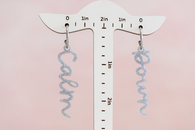 May include: A pair of silver glitter earrings with the names "Carolyn" and "Rowan" hanging from a ruler measuring 2 inches.