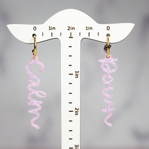 May include: A pair of pink glitter hoop earrings with the names "Caryn" and "Kevin" hanging from them. The earrings are displayed on a white ruler with inch markings.