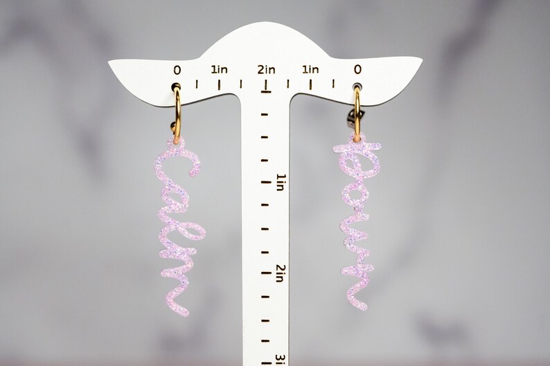 May include: A pair of pink glitter hoop earrings with the names "Caryn" and "Kevin" hanging from them. The earrings are displayed on a white ruler with inch markings.