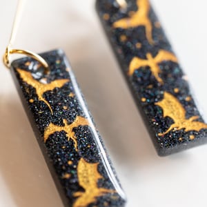 May include: A pair of black and gold earrings with a glittery finish. The earrings are rectangular in shape and feature a repeating pattern of gold bats.