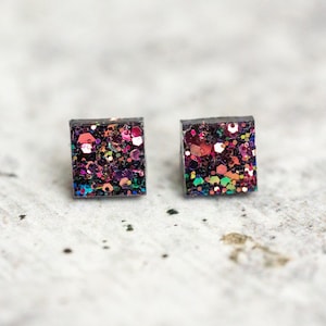 May include: A pair of square-shaped stud earrings with a black base and a colorful glitter design. The glitter is a mix of pink, purple, green, and gold.