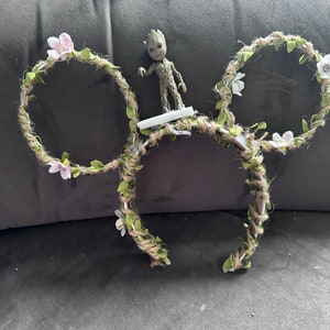 May include: A pair of brown and green Mickey Mouse ears with white flowers and leaves. A small green figurine of a character from the movie Guardians of the Galaxy is standing on top of the ears.