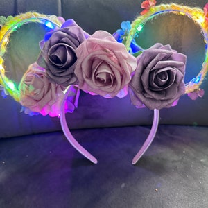Puede incluir: Flores de tela moradas, rosas y blancas adornan un par de orejas de ratón iluminadas. Las orejas están hechas de un material tejido y tienen una diadema blanca.