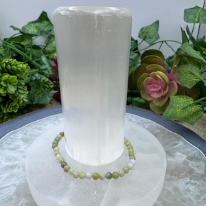 May include: A light green beaded bracelet rests on a white, cylindrical selenite crystal. The bracelet features small, round beads in shades of green and cream. The selenite crystal is displayed on a white, circular base.