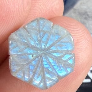 May include: A hexagonal labradorite gemstone carved with a radial starburst pattern, displaying iridescent blue flashes. The stone is positioned next to a black ruler showing metric markings, with the visible portion starting at the 13 centimeter mark.