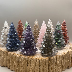 May include: A collection of gemstone Christmas tree figurines in various colors, including clear, blue, brown, green, purple, pink, and gray. The trees are arranged on a rustic, textured wooden base, showcasing their detailed, layered design. The background is a neutral gray.