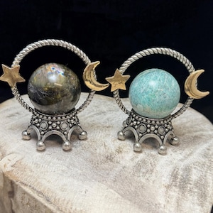 May include: Two decorative crystal ball holders. Each has a silver-colored twisted ring with a gold star and crescent moon accent. One holds a green-grey sphere, the other a blue-green sphere. The bases are silver-colored with a crown-like design.