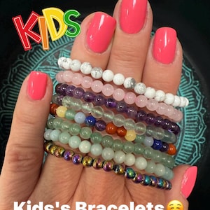 May include: A collection of beaded bracelets in various colors, including white, pink, purple, green, and iridescent, held in a hand with bright pink nail polish. The image includes the text "KIDS" and "Kids's Bracelets."