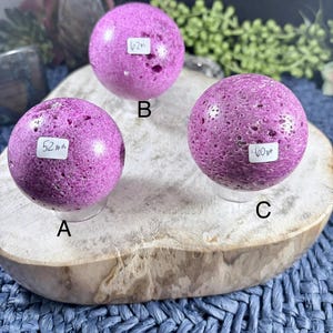May include: Three spherical, textured, purple gemstones are displayed on a wooden slab. Each sphere has a unique size, indicated by a label: 52mm, 62mm, and 60mm. The gemstones are set against a backdrop of greenery and a blue woven surface.
