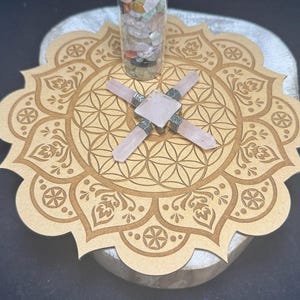 May include: A clear glass bottle filled with colorful gemstones, topped with a cork, sits on a wooden mandala. A rose quartz crystal grid is centered on the mandala, with four crystals extending outwards.