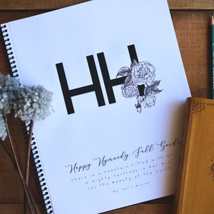 May include: A white spiral bound notebook with the text "Happy Handmade Fall Guide" and the letters "HH" in black with a floral design. The notebook is on a wooden surface with gray yarn pom-poms and colored pencils.