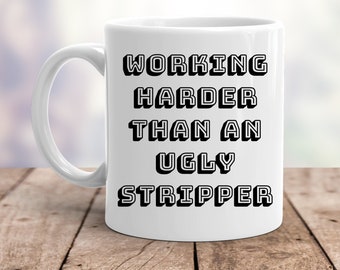 May include: White ceramic mug with the text "WORKING HARDER THAN AN UGLY STRIPPER" in black, block letters. The mug is on a wooden surface with a blurred background.