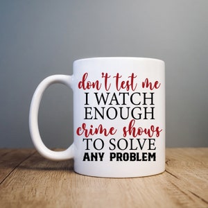 以下が含まれることがあります： 黒と赤のデザインが施された白いセラミック製のマグカップ。マグカップには「don't test me I watch enough crime shows to solve any problem」という文字が書かれています。