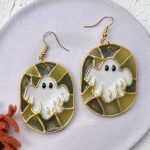 Może przedstawiać: Para złotych kolczyków na Halloween. Każdy kolczyk ma biały wzór ducha z czarnymi oczami, osadzony w oliwkowozielonej i złotej ramie. Kolczyki są prezentowane na jasnofioletowym talerzu, z pomarańczową przędzą w tle.