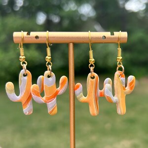 Könnte beinhalten: Vier Paar Ohrringe in Kaktusform. Die Ohrringe bestehen aus marmoriertem Polymer Clay in Orange, Gelb und Grau. Sie hängen an goldfarbenen Haken. Die Ohrringe werden auf einem goldfarbenen Ohrringständer präsentiert.