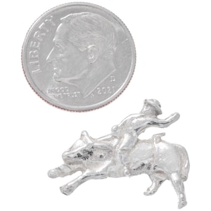 May include: A silver charm depicting a cowboy riding a bucking bronco. The charm is about the size of a dime.