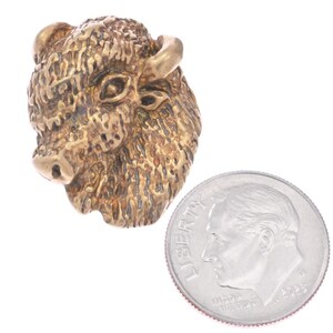 May include: A bronze-coloured buffalo head sculpture with detailed fur and prominent features, including horns and a snout. A UK coin is next to the sculpture for size comparison.