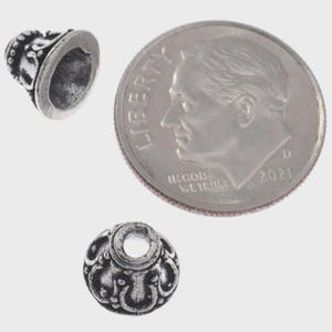 May include: Close-up of two silver-colored metal beads with ornate designs, next to a U.S. dime coin. The beads have a cone shape and are likely used for jewelry making or crafts. The dime provides a size reference.