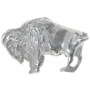 May include: A silver metal figurine of a bison, standing on all four legs with its head lowered. The figurine is detailed with textured fur and a rough, slightly uneven surface.