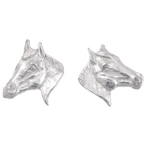 May include: A pair of silver-colored stud earrings, each shaped like a horse's head. The earrings are detailed with the features of the horse's face, including the eyes, nose, and ears. The earrings are set against a white background.