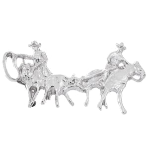 May include: A silver-toned metal brooch depicting two cowboys on horseback roping a steer. The brooch has a detailed, three-dimensional design, showcasing the figures and their horses in action. The brooch is a Western-themed accessory.