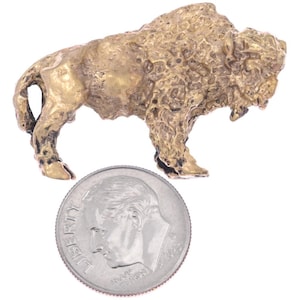 May include: A small, detailed brass figurine of a bison, standing with its head lowered. The bison has a textured surface and is shown next to a coin for size comparison. The figurine is a miniature sculpture.