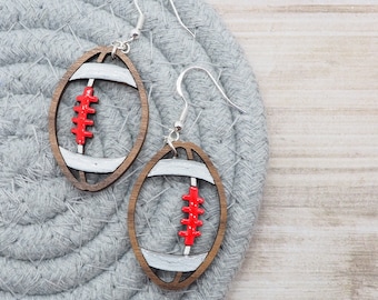 May include: A pair of wooden earrings shaped like footballs. The footballs are painted white with red stitching and a silver outline. The earrings are hanging from silver hooks.
