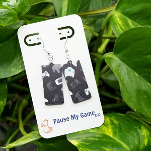 May include: A pair of silver dangle earrings with black and white rectangular charms featuring a pattern of video game controllers and consoles. The charms are attached to silver hooks. The earrings are displayed on a white card with the text "Pause My Game LLC".