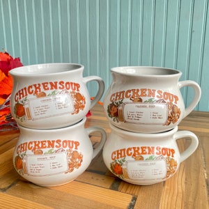 May include: Four white ceramic soup mugs with handles, each featuring the words "CHICKEN SOUP" in orange lettering. The mugs have a vintage design with a recipe graphic. The mugs are stacked on a wooden surface.