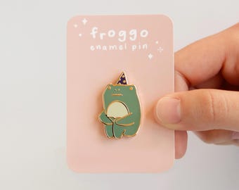 May include: A green frog enamel pin wearing a dark blue party hat with gold stars, attached to a light pink rectangular display card. The card features the white text "froggo enamel pin" at the top, accented with small white star graphics.
