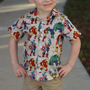 May include: A short-sleeved button-down shirt with a beige background and a colorful print of various superheroes, including Captain America, Iron Man, and Hulk.