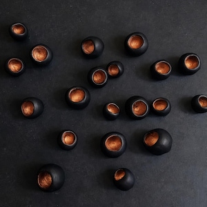 Puede incluir: Disposición de objetos esféricos negros con interior metálico color cobre. Los objetos están dispersos sobre un fondo oscuro, creando un patrón visualmente interesante. Las esferas varían en tamaño.